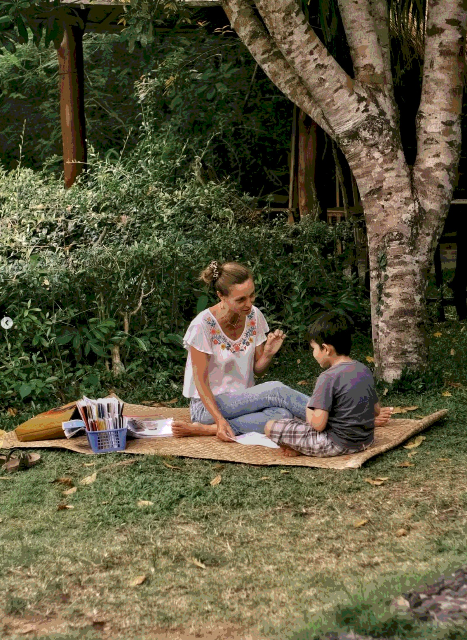 A teacher reading with a student outdoors on a mat under a tree at Wisdom Academy